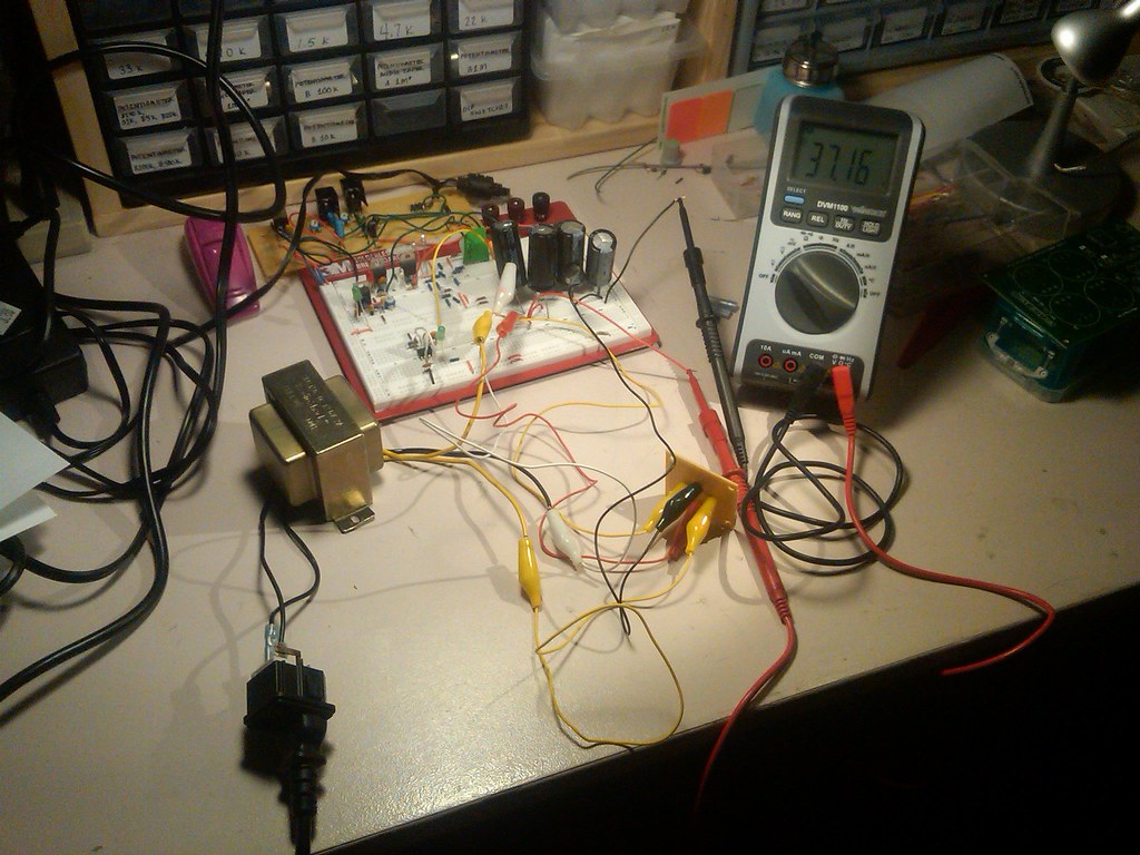 Figure 3 — 3 — A low-voltage supply on the bench: a small mains transformer (lower left) feeding a board of through-hole parts — diodes, electrolytic capacitors, transistors — with a multimeter readi…